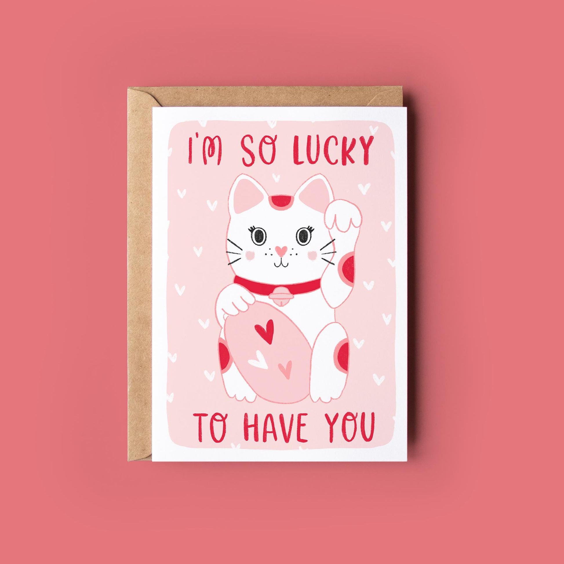 May include: A greeting card with a white lucky cat illustration with red spots, holding a pink oval with hearts. The text "I'M SO LUCKY TO HAVE YOU" is in red. The card is in a brown envelope, on a pink background.