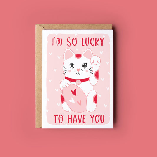 May include: A greeting card with a white lucky cat illustration with red spots, holding a pink oval with hearts. The text "I'M SO LUCKY TO HAVE YOU" is in red. The card is in a brown envelope, on a pink background.